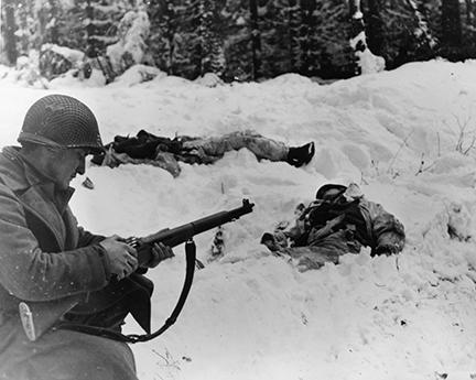 Ardennes-Battle of the Bulge. January 15, 1945 - Pfc Frank Vukasin of Great Falls, Montana, stops to load clip in rifle while advancing in snow-covered front line sector at Houffalize, Belgium. In background, two dead German soldiers are wearing camouflage snow suits.