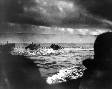 Coast Guard landing barges hitting the French coast with the first wave of the invasion. American soldiers wade ashore under heavy machine gun fire from Nazi beach nests.
