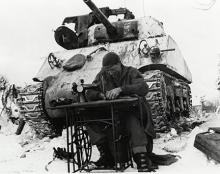 Ardennes-Battle of the Bulge. January 23, 1945 - Sgt. William Phelps of San Antonio, Texas, a tanker of the 42nd Tank Battalion, mends his combat clothes in Steinbach, Belgium. He uses a sewing machine in front of tank and snow covers the ground.